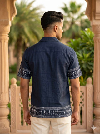 A man stands with his back to the camera, wearing the Koyra Folkline Shirt—a navy relaxed short-sleeve shirt featuring white tribal patterns on the sleeves and hem—paired with cream pants. He is outdoors near stone pillars and lush green foliage.