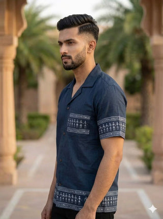 A man with short dark hair and a beard stands outdoors, wearing the Koyra Folkline Shirt—a relaxed short-sleeve shirt featuring white tribal patterns. Palm trees and a stone structure are visible in the background.