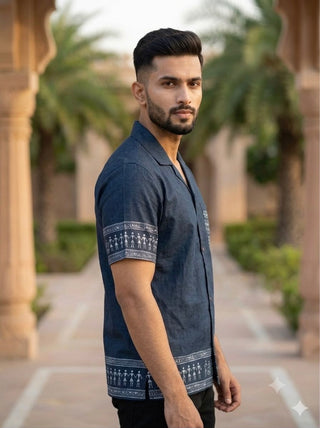 A man with short hair and a beard stands outdoors, wearing the Koyra Folkline Shirt—a relaxed short-sleeve style in blue with white patterns. Palm trees and stone columns are visible in the background.