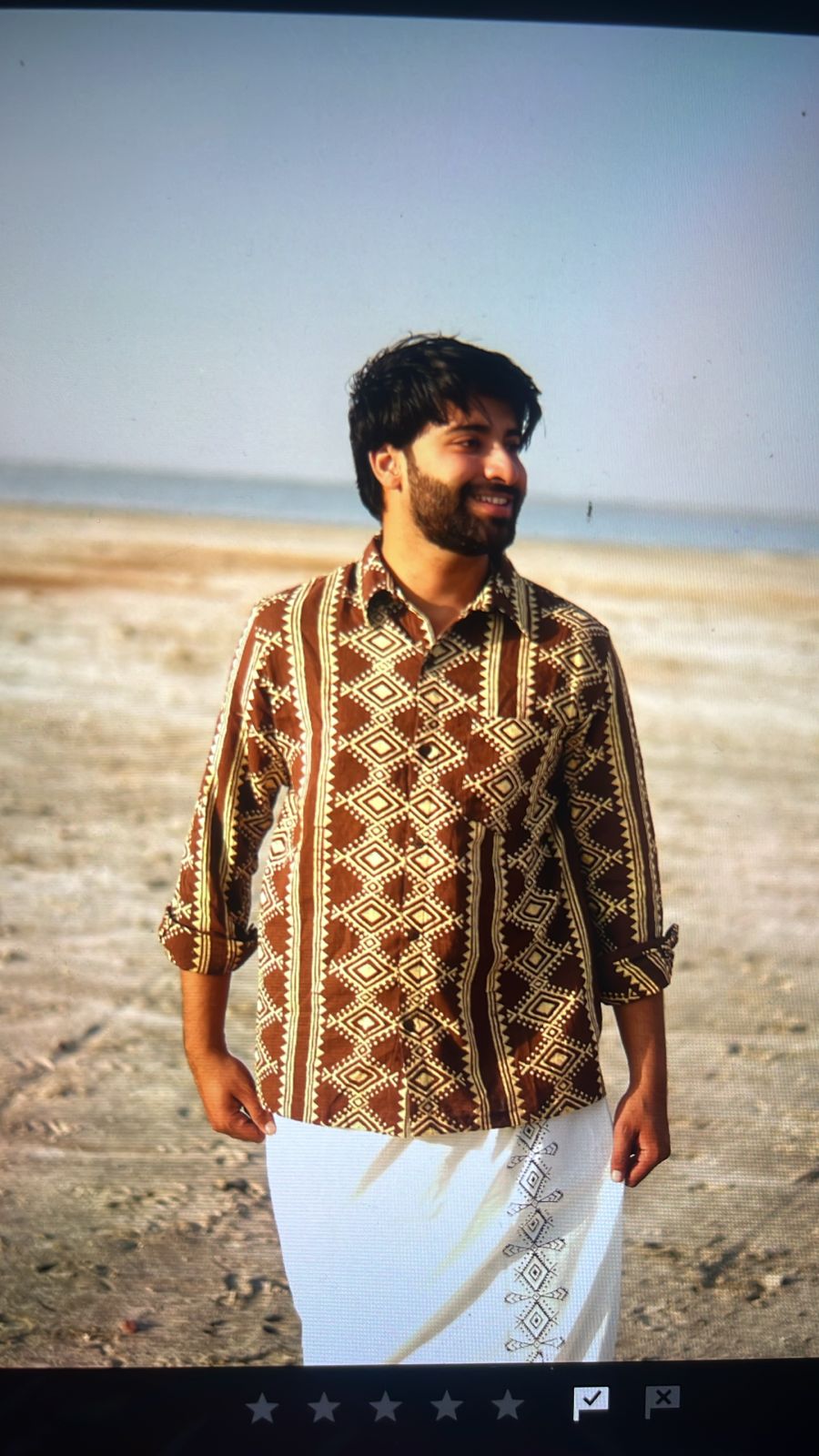 A man with dark hair and a beard stands smiling on a sandy beach, sea and sky in the background, wearing the Varun Handblock Printed Cotton Shirt—a testament to Indian craftsmanship—paired with a white traditional wrap.