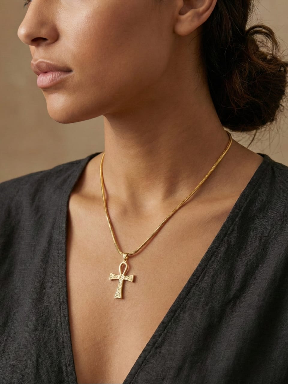 A person shown from the shoulders up against a neutral background wears a black V-neck top and the Amun Life Key Necklace, featuring a gold Ankh pendant.