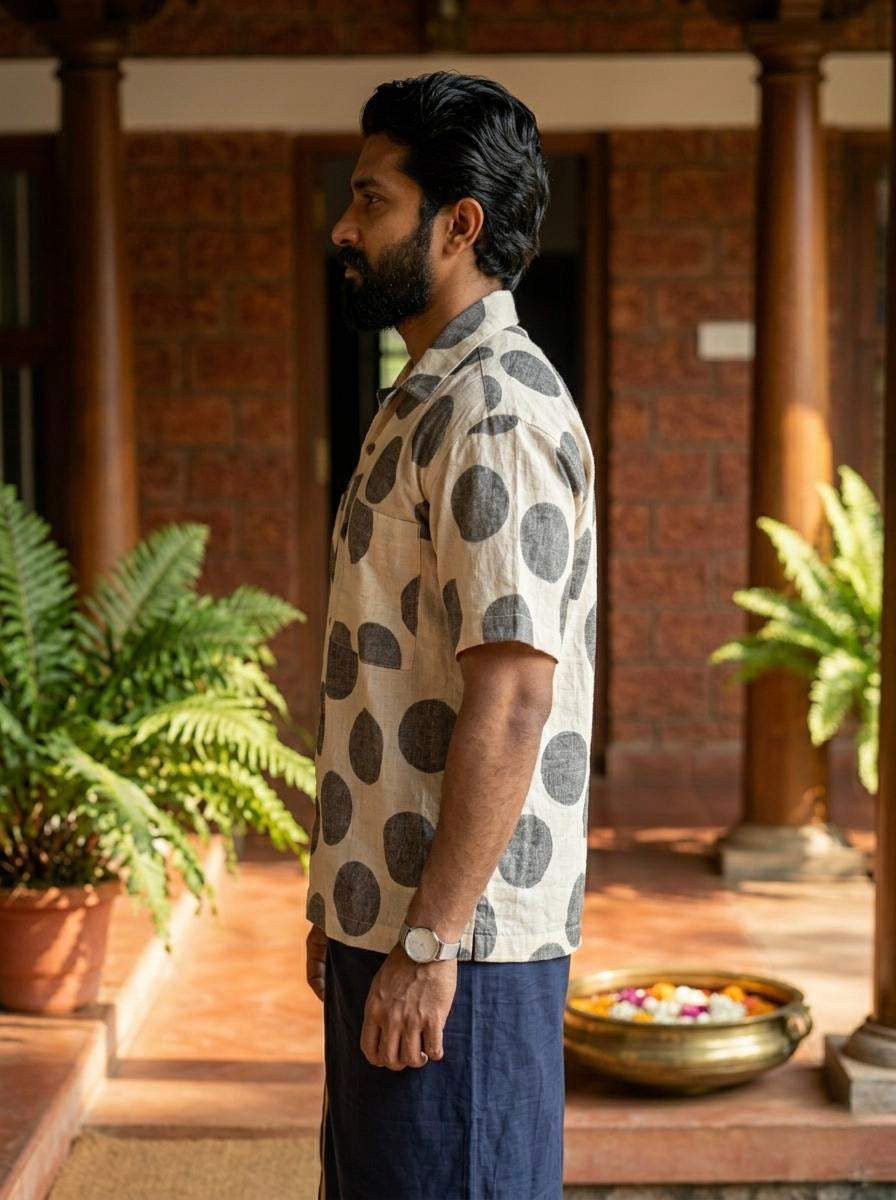A man with a beard stands in profile, wearing the Dot Breeze Shirt—a beige half-sleeve shirt featuring oversized dot patterns and crafted from breathable lightweight fabric—paired with navy shorts. He is indoors, beside potted ferns and a brass bowl of floating flowers, set against wooden pillars and brick walls in the background.