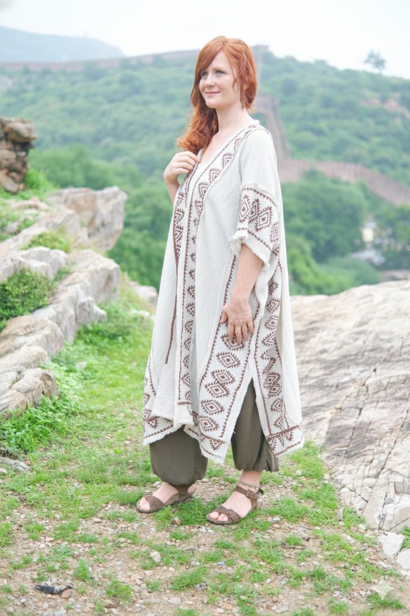 Full-length front view of the model in the natural cream knit kimono with the brown block printed border and cobweb fringe hem, worn open over olive harem pants with sandals, on a hillside path with green mountains behind.