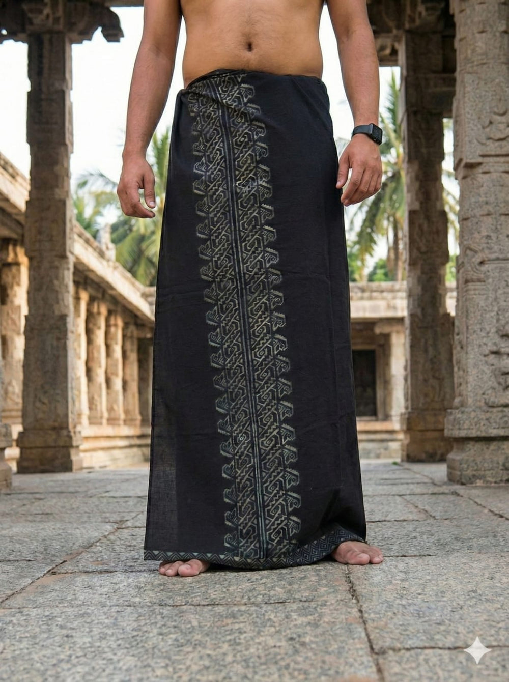 A bare-chested person stands in a stone corridor, wearing the Sanga Dhoti in black with intricate gold patterns inspired by tribal handblock print, a watch on their wrist, and no shoes. Their face is not visible.