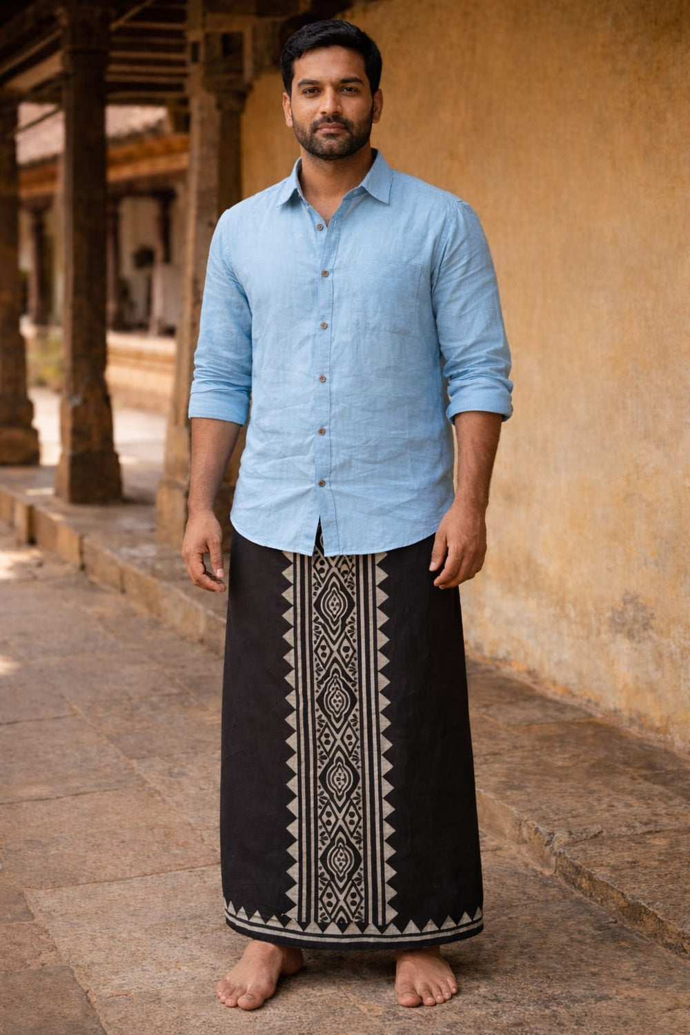 A man stands barefoot on stone pavement, wearing a light blue shirt and the Jasper Dhoti—black with geometric block print patterns—against a rustic wall and pillars.