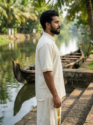 Wearing the Midnight Nomad kurta, a man stands beside a river lined with wooden boats and lush green palm trees, bathed in soft daylight. The traditional outfit features delicate handblock prints in a creamy hue.