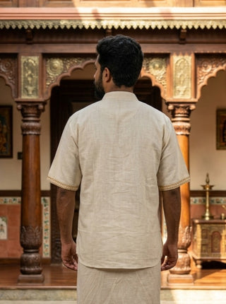 A man wearing the Midnight Nomad kurta—a beige short-sleeved traditional outfit featuring subtle handblock prints—stands with his back to the camera in front of an ornate wooden structure adorned with columns and intricate carvings.