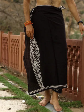 A person in a black Tribal Dhoti featuring white spiral designs, reminiscent of traditional Indian style, stands barefoot on a path beside a decorative railing. The background shows trees and grass, with the upper body partially hidden.