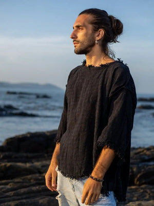 Black baggy knit T-shirt side view at the shoreline, showing the longer back hem and frayed sleeve edge
