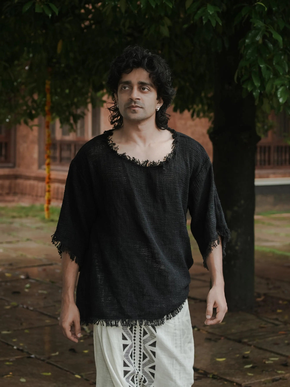 Black cotton-jute baggy knit T-shirt on a model walking under trees, frayed neck and sleeve hems, pale trousers