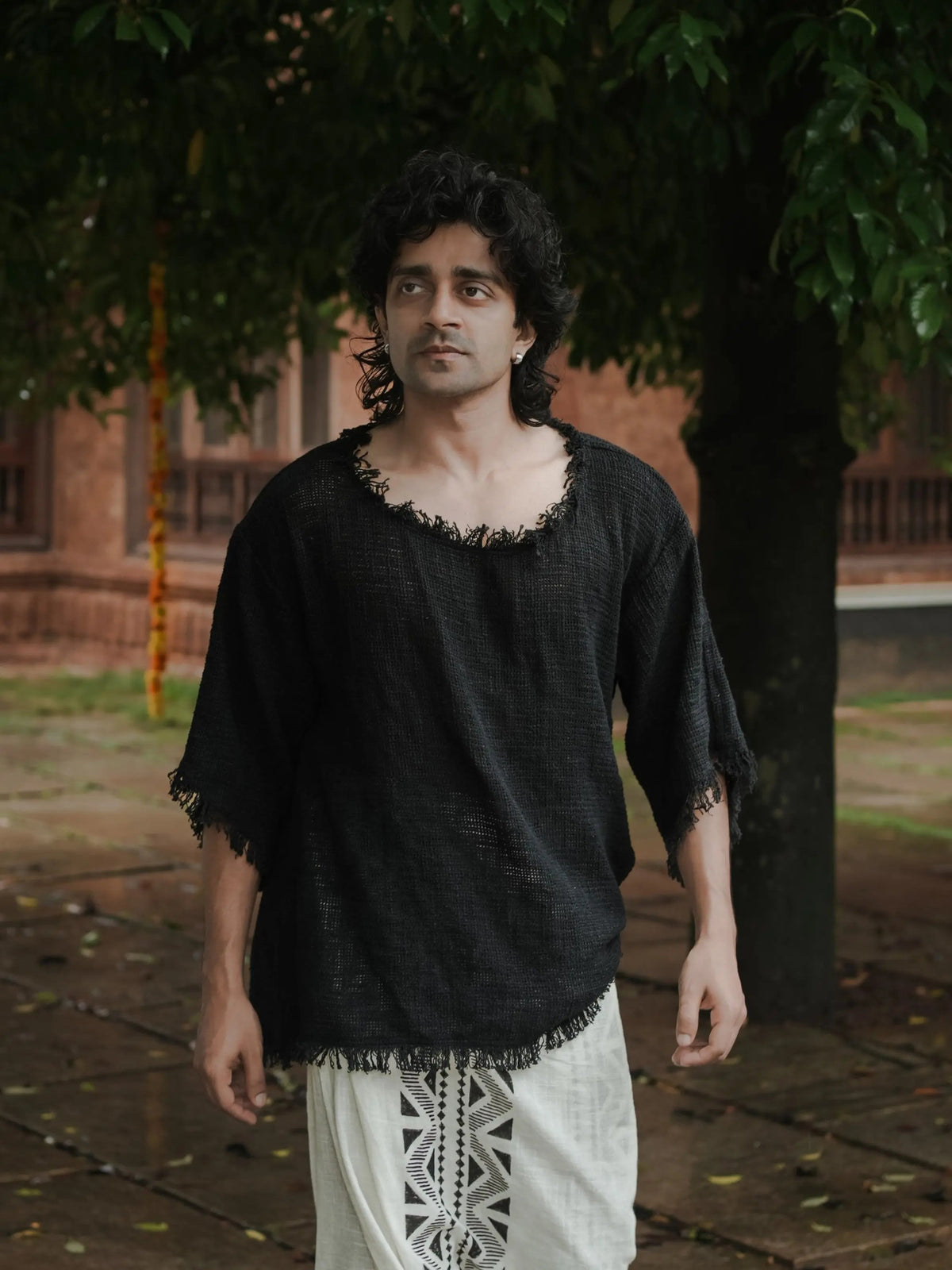 Black cotton-jute baggy knit T-shirt on a model walking under trees, frayed neck and sleeve hems, pale trousers