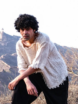 Natural cream frayed-edge jute T-shirt on a model crouching on rocky ground, open-weave texture in sunlight