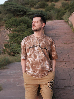 Front view of the Become the Light brown tie-dye oversized cotton tee worn on a stone path, chest print visible.