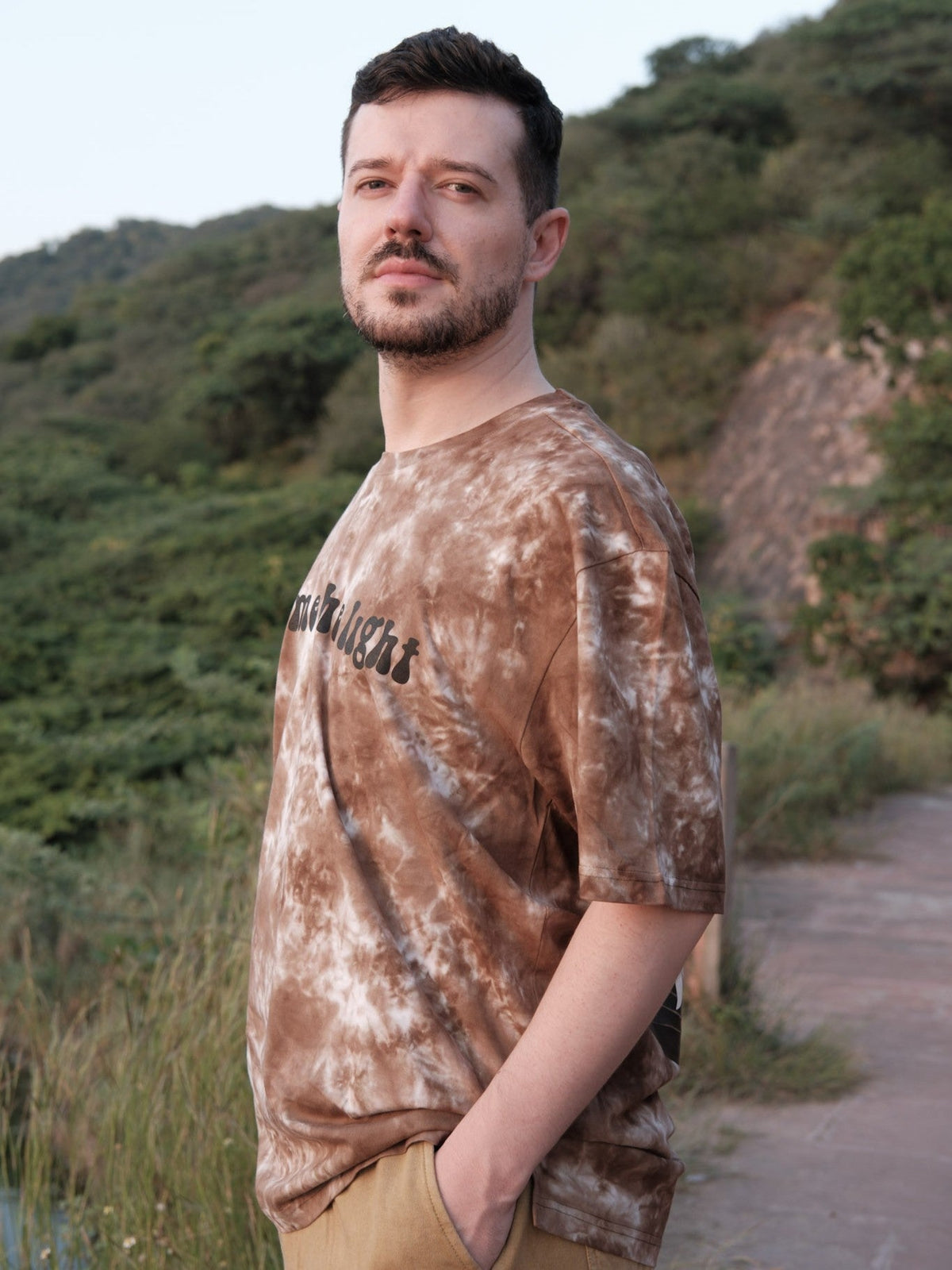 Three-quarter view of the Become the Light front print on a brown tie-dye oversized cotton tee against green hills.
