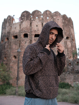 Black maze-print hoodie worn hood up, close crop of male model holding hood, fort tower behind