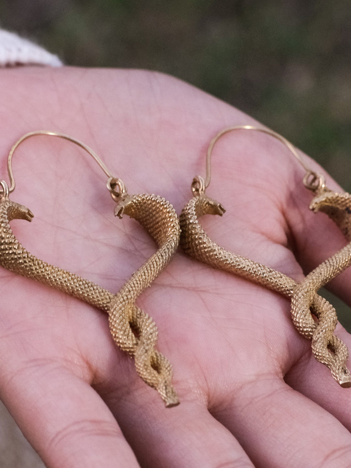 Both Brahman brass serpent drop earrings held in an open palm, showing the full intertwined snake design and textured finish