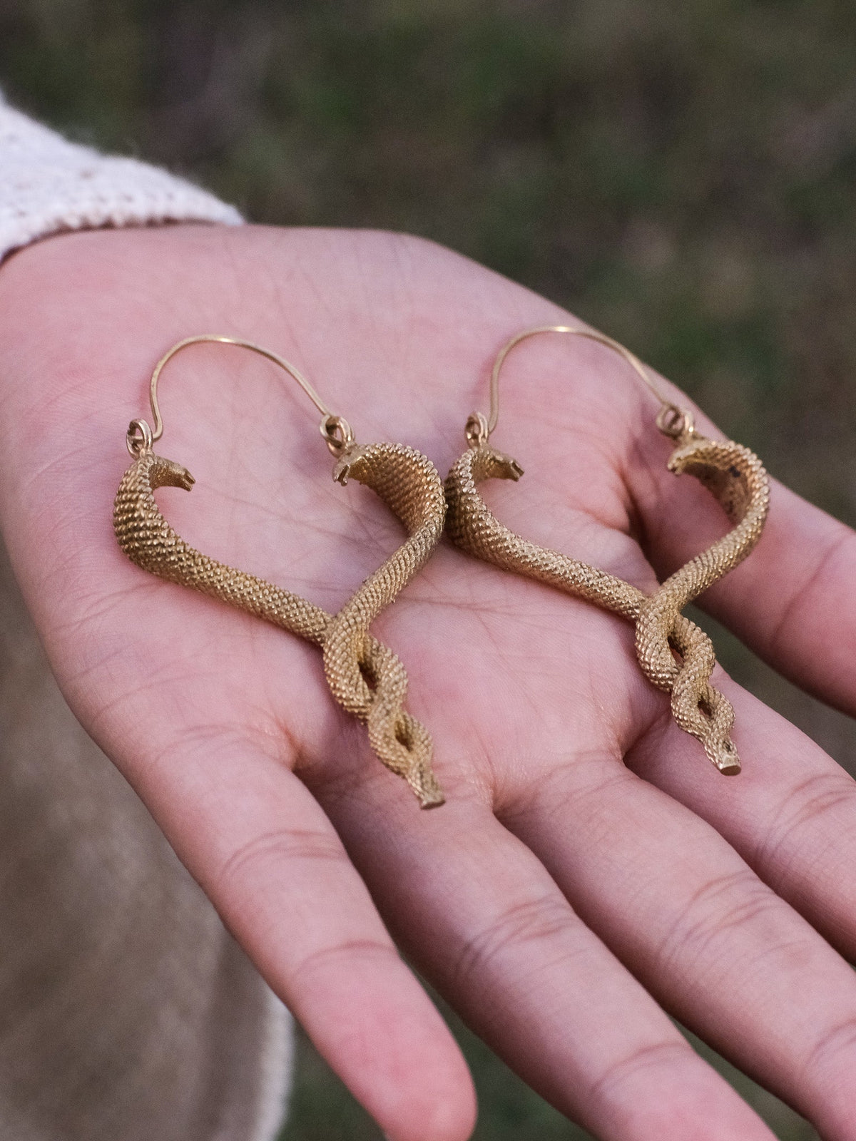 Both Brahman brass serpent drop earrings laid in an open palm against a blurred outdoor backdrop, showing the matched pair