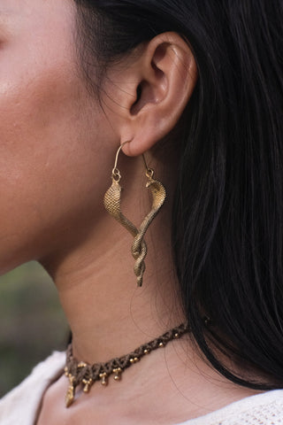 Close-up of the Brahman brass serpent drop earring worn on a model's ear, showing the textured antique-gold finish outdoors