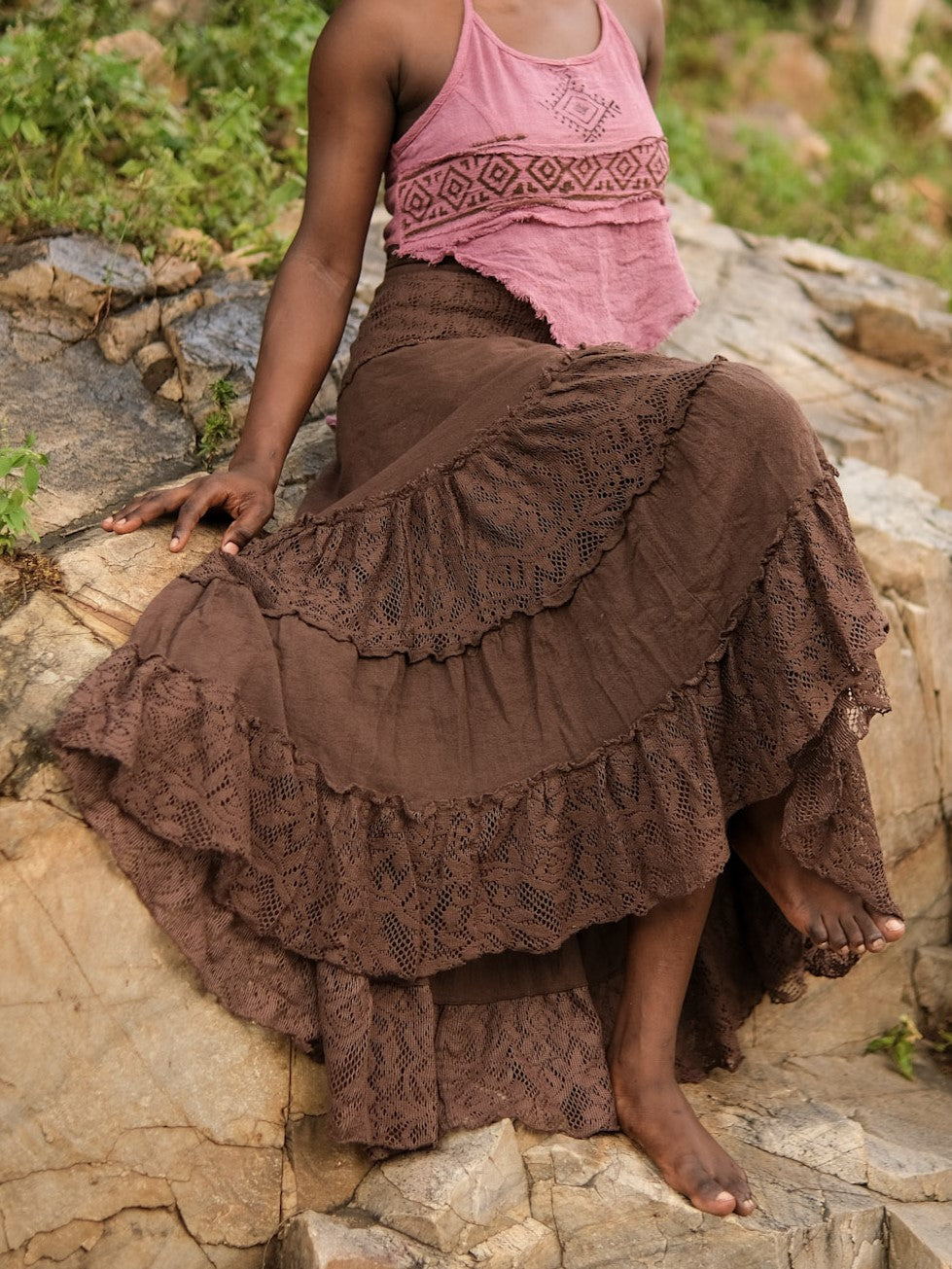 Seated model in a brown khadi cotton wrap skirt on grey rocks, openwork lace tier detail across the thigh