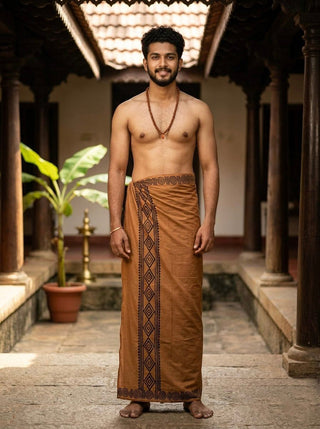 Man in full-length Camel Brown Ory Dhoti with hand-printed border, standing barefoot in a traditional wooden corridor