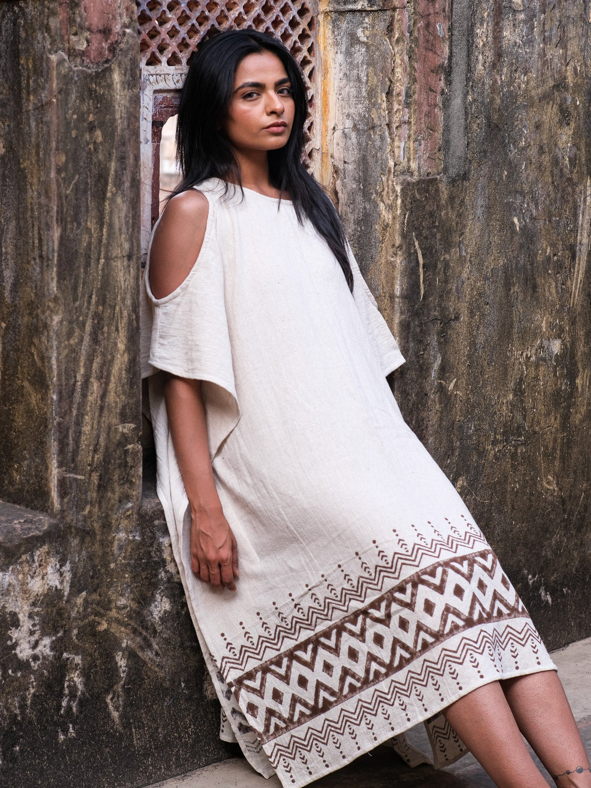 Chandra Kaftan side view, model seated against weathered stone wall, cream kaftan with brown block print border