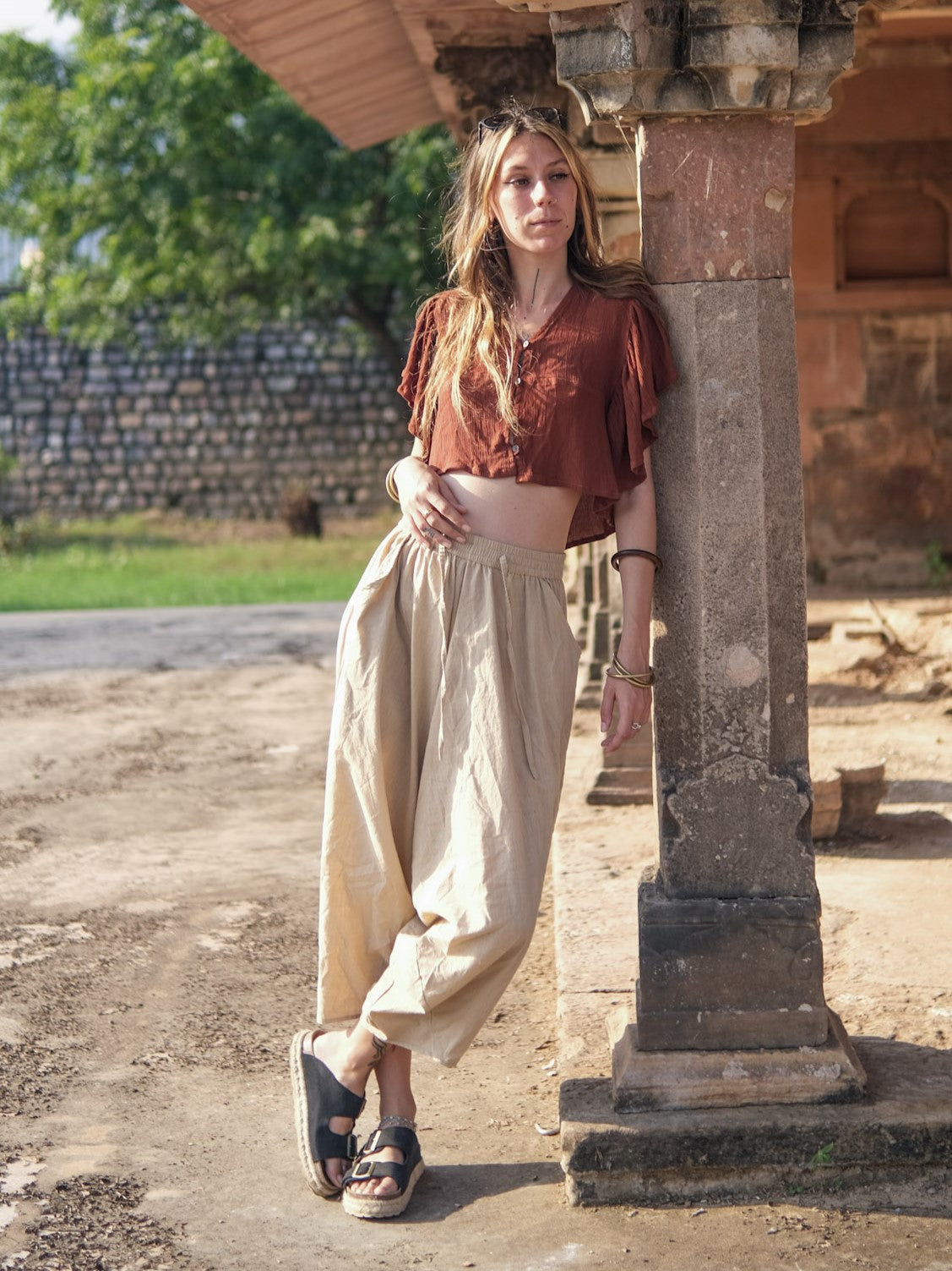 Beige cotton harem pants worn with a rust blouse and sandals, leaning on a carved sandstone pillar in warm daylight.