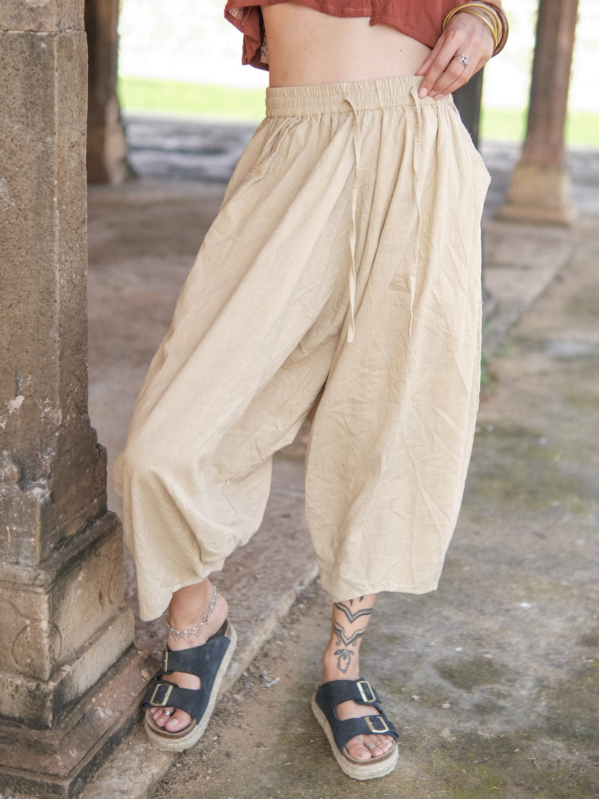 Warm beige cotton sirwal pants worn with anklets and sandals beside a carved temple pillar, wide leg gathering at the ankle.