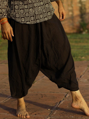 Black sirwal harem pants walking barefoot through a stone courtyard with a printed top, the leg gathered at the ankle.