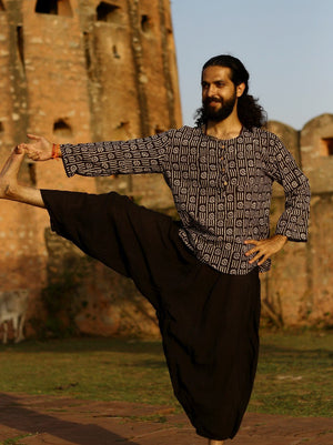 Black cotton harem pants worn by a man in a printed shirt lifting one leg in a balance pose at sandstone fort ruins.