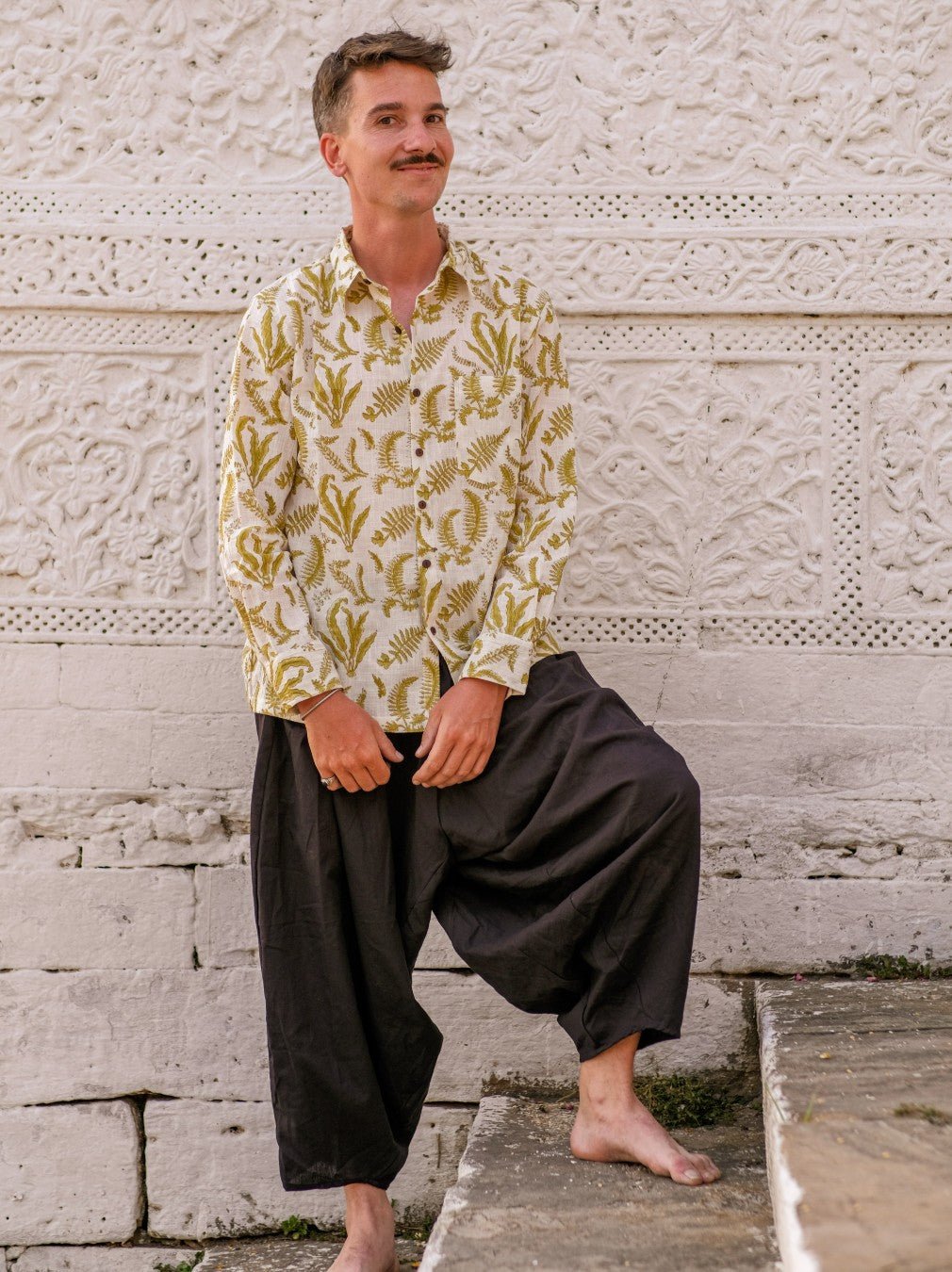 Black cotton harem pants worn with a leaf print shirt, standing barefoot against a carved white stone wall.