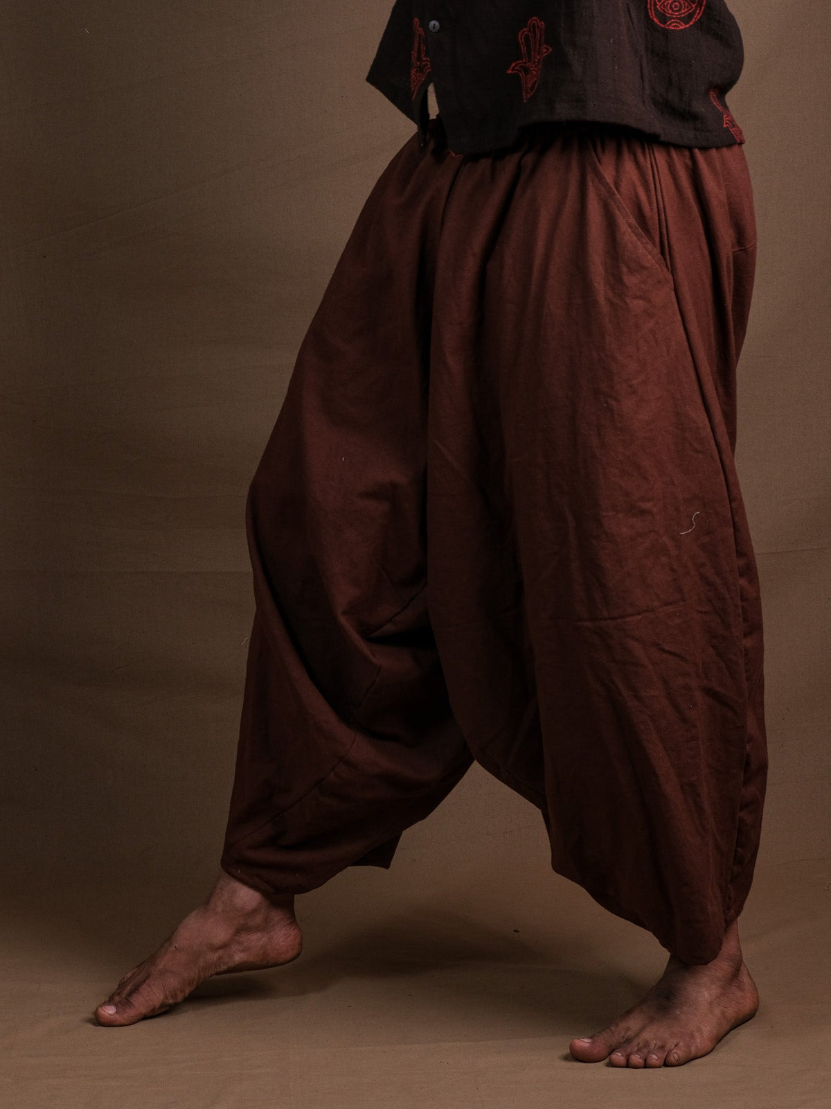 Rust brown harem pants photographed against a plain brown studio backdrop, walking pose showing the deep drop crotch.