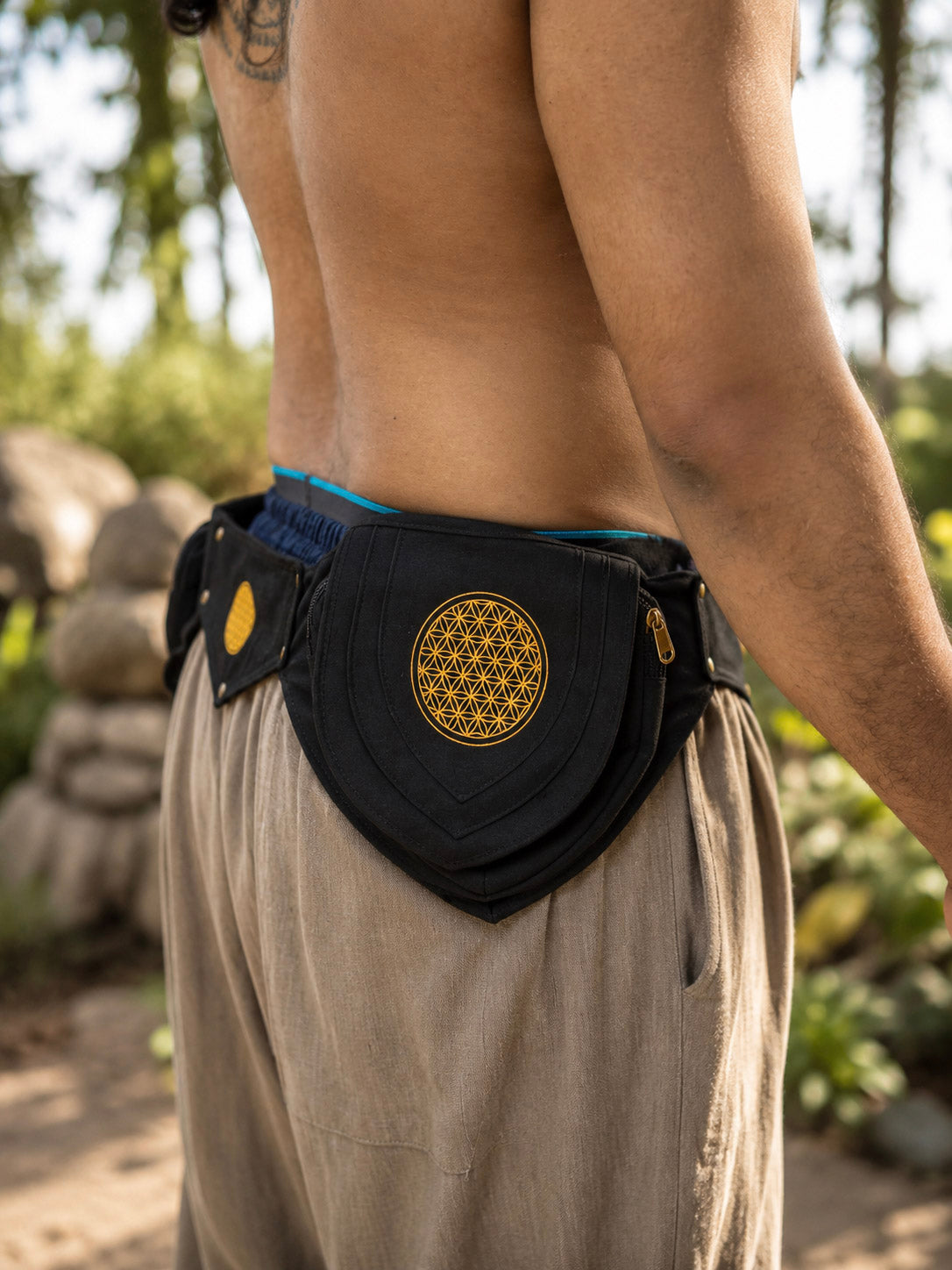 Wearing loose beige pants and the Mystic Waist Belt—a black fabric festival accessory featuring a yellow geometric design—a person stands outdoors amid greenery and rocks.