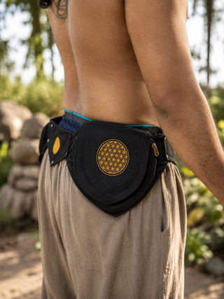Wearing loose beige pants and the Mystic Waist Belt—a black fabric festival accessory featuring a yellow geometric design—a person stands outdoors amid greenery and rocks.