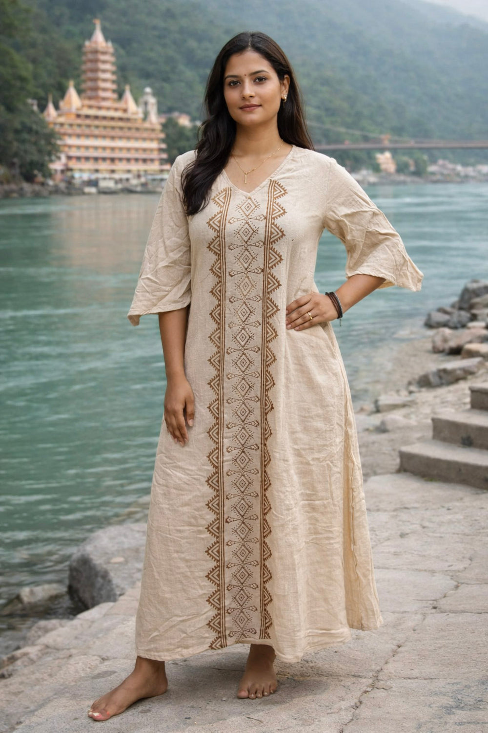 A woman in the beige Garga Dress stands barefoot by a river, framed by mountains, stone steps, and a large temple in the background. The elegant Garga Dress features subtle block printing that harmonizes with the serene landscape.