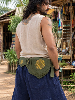 A man with long hair and a tattoo wears a sleeveless beige shirt, blue pants, and the Mystic Waist Belt in green. Market stalls with crafts are visible in the background.