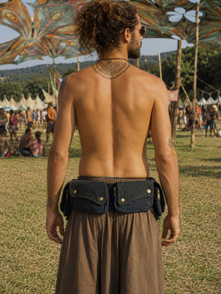 A person with curly hair in a bun stands shirtless at an outdoor festival, wearing loose brown pants and accessorizing with the Rustic Festival Bag—a black utility belt bag. Tents and people are visible in the background beneath a colorful canopy, enhancing the bohemian style atmosphere.