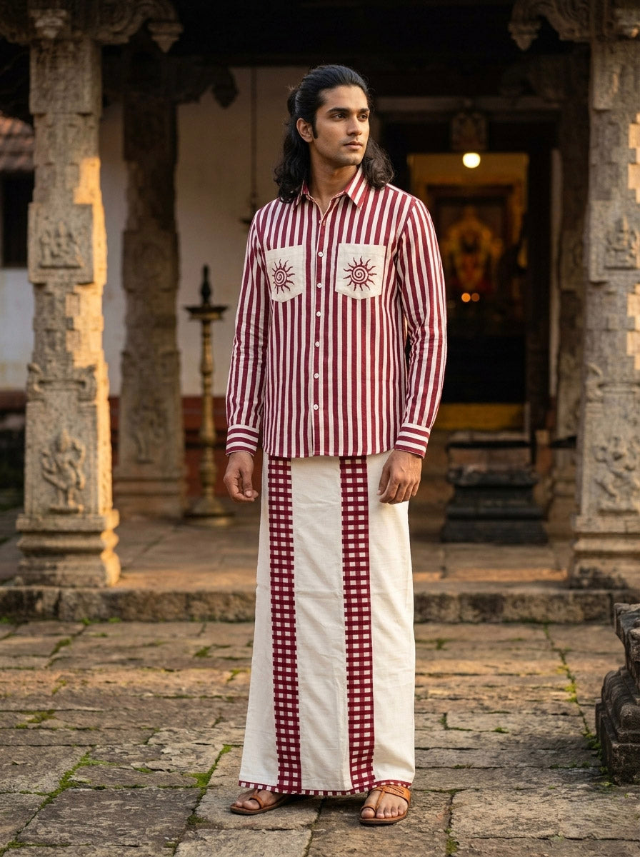 A man wearing the maroon Surya Rekha Shirt, featuring artisan sun designs and stripes, paired with a matching patterned dhoti, stands in front of an ancient stone temple.