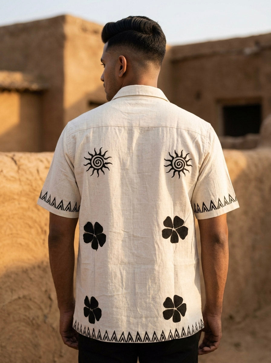 A man with short dark hair stands outdoors facing away, wearing the Kovah Sun Shirt—handcrafted in beige with black sun and clover designs on the back, and geometric patterns along the sleeves and hem. Adobe-style buildings are visible in the background.