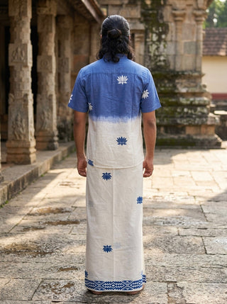 A person stands outdoors, facing away, wearing the Sarovar Shirt—characterized by its blue-to-ivory ombre and lotus motif—and paired with a coordinating traditional white garment decorated with blue patterns, set against ancient stone architecture.