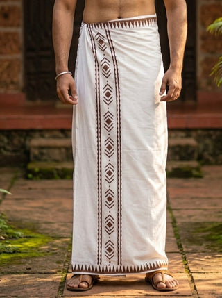 A man stands outdoors wearing the Mehfil Dhoti, which features traditional cotton fabric and brown geometric patterns along the border, adding an ethnic touch to his look; his upper body is bare, and he pairs the outfit with sandals.