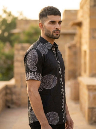 A man with short hair and a beard stands outdoors, wearing the Obsid Shirt—a black cotton shirt adorned with white paisley designs. In the softly illuminated background, blurred stone buildings and greenery can be seen.
