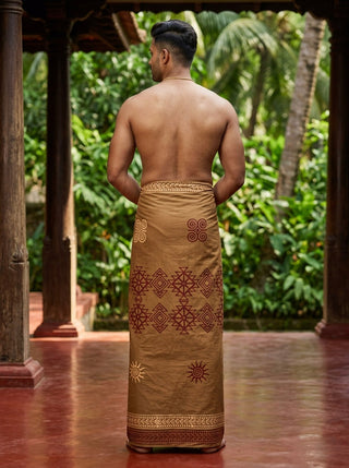 A man stands barefoot with his back to the camera, wearing the Ramya Dhoti—a brown, patterned hand block print garment—wrapped around his waist. He is shirtless, with lush green plants and trees visible in the background.
