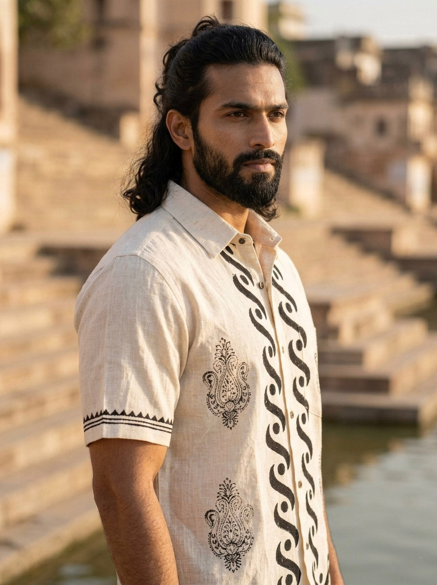 A man with long dark hair and a beard stands outdoors near stone steps and water, wearing the Elio Shirt—a light-colored short-sleeve artisan shirt featuring black traditional patterns.