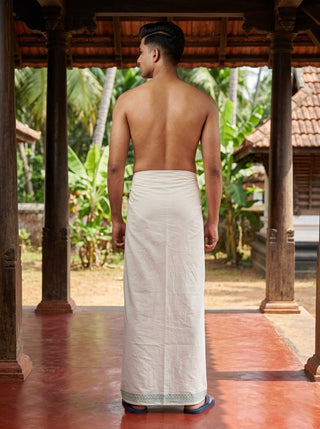 A man stands barefoot on a veranda, facing away, wearing the traditional white unisex Naaro Dhoti made of cotton. Lush green plants and wooden pillars surround him, with tiled roofs and palm trees in the background, evoking the classic elegance of South Indian mundu—making it perfect for resort beach wear.
