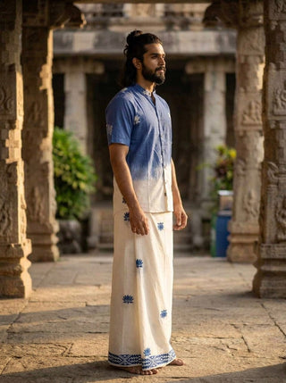 A man with a beard and long hair tied back stands barefoot in a traditional stone corridor, wearing the Sarovar Shirt paired with a white dhoti adorned with blue lotus motifs. He gazes to the side as soft sunlight falls across him.