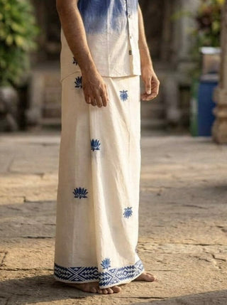 A person stands barefoot on stone ground, wearing the cream-colored Jalaj Dhoti—a traditional wrap-around mundu adorned with blue handcrafted lotus motifs and paired with a matching shirt. The head is not visible in the image.