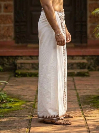 A man standing outdoors wears the Mehfil Dhoti, featuring a decorative border, paired with brown sandals and a bracelet for an ethnic touch. The background includes stone steps and a building entrance.