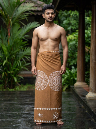 A man stands barefoot outdoors on wet stone, dressed in the Kavu Dhoti—a brown heritage-inspired garment featuring white geometric block print patterns—with greenery and wooden pillars in the background.