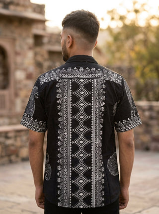 A man stands outdoors facing away, wearing the Obsid Shirt—a black cotton shirt featuring intricate white geometric and paisley patterns. Stone architecture and trees appear in the softly blurred background.