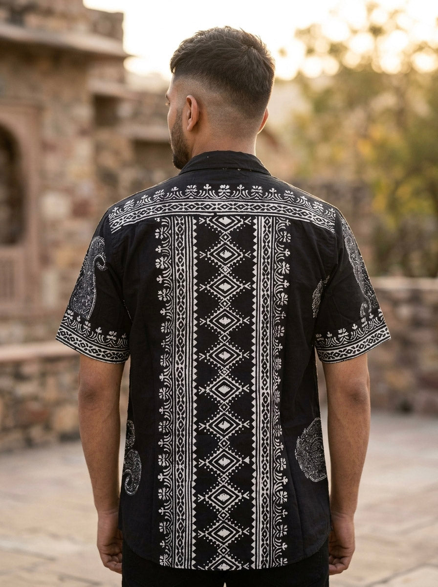 A man stands outdoors facing away, wearing the Obsid Shirt—a black cotton shirt featuring intricate white geometric and paisley patterns. Stone architecture and trees appear in the softly blurred background.